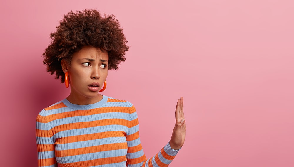 Discontent African American woman keeps palm in refusal gesture, says stays away from me and do not come closer, avoids crowded places, grimaces disappointed, isolated on pink background, empty space